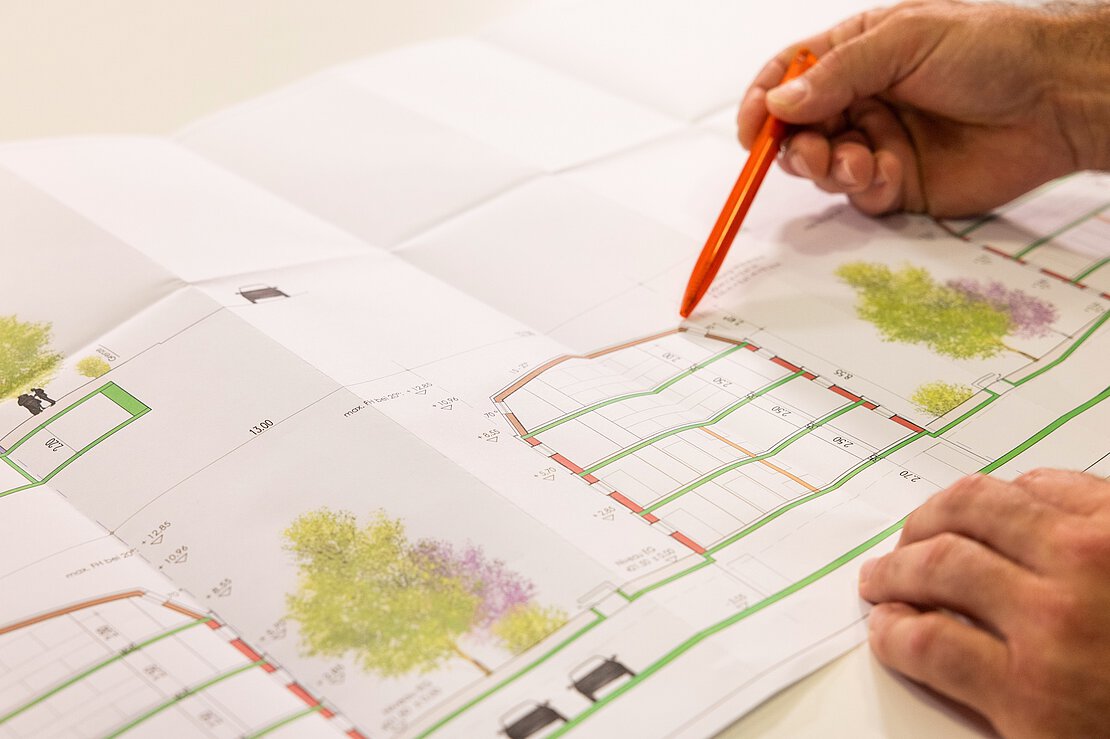 Detailarbeit am Bauplan Nahaufnahme einer Hand mit orangefarbenem Stift, die auf einen detaillierten Bauplan mit Grünflächen zeigt.